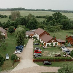 Piotr Klentak - Panoramiczny widok z lotu ptaka na Dom Weselny z zaparkowanymi samochodami i gośćmi w otoczeniu zielonych pól i drzew.