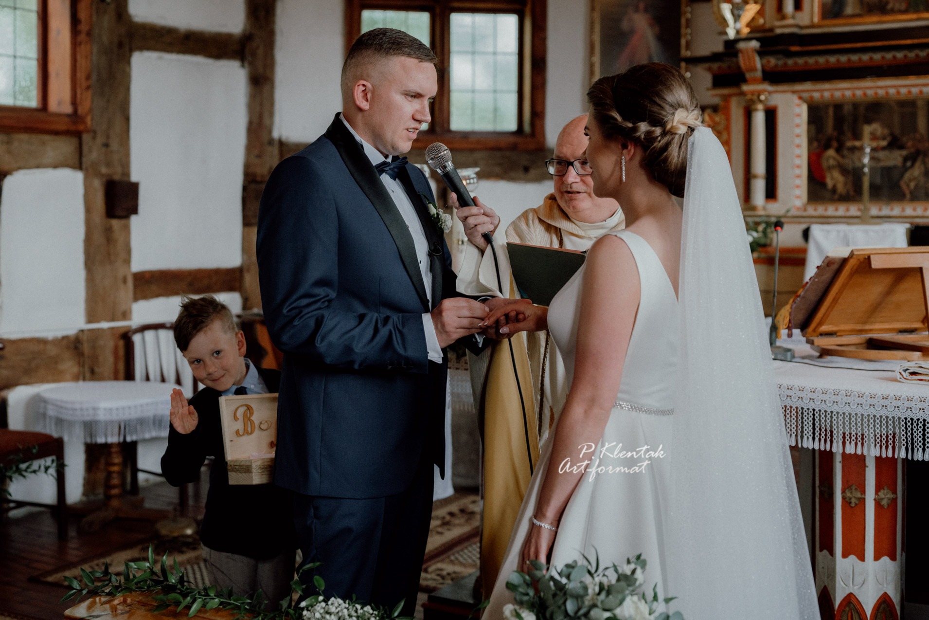 Para młoda podczas ceremonii ślubnej w drewnianym kościele, wymiana obrączek, w tle ministrant z pudełkiem na obrączki i ksiądz.