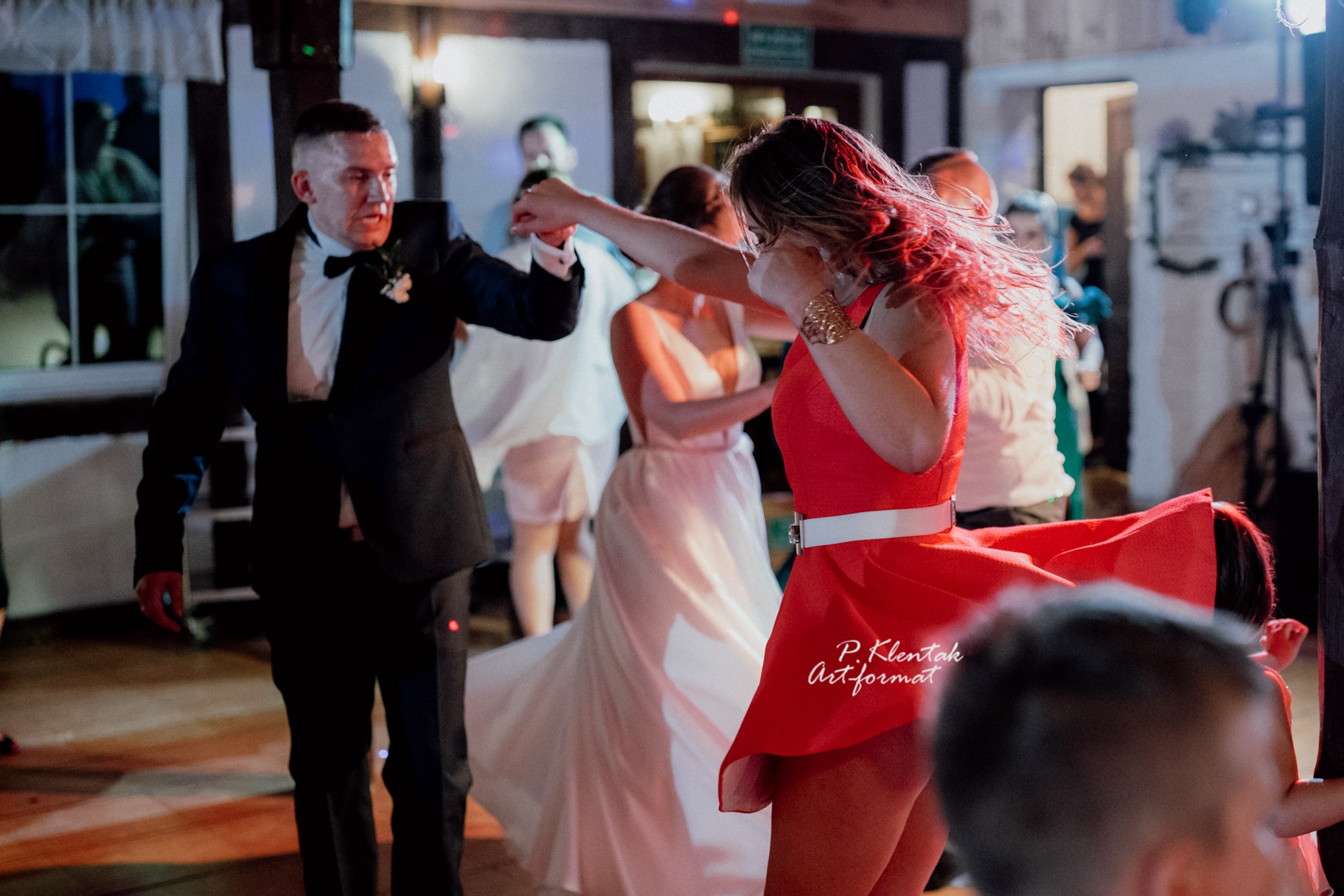 Goście weselni tańczą podczas przyjęcia, panna młoda w białej sukni w tle, kobieta w czerwonej sukience z białym paskiem na pierwszym planie.