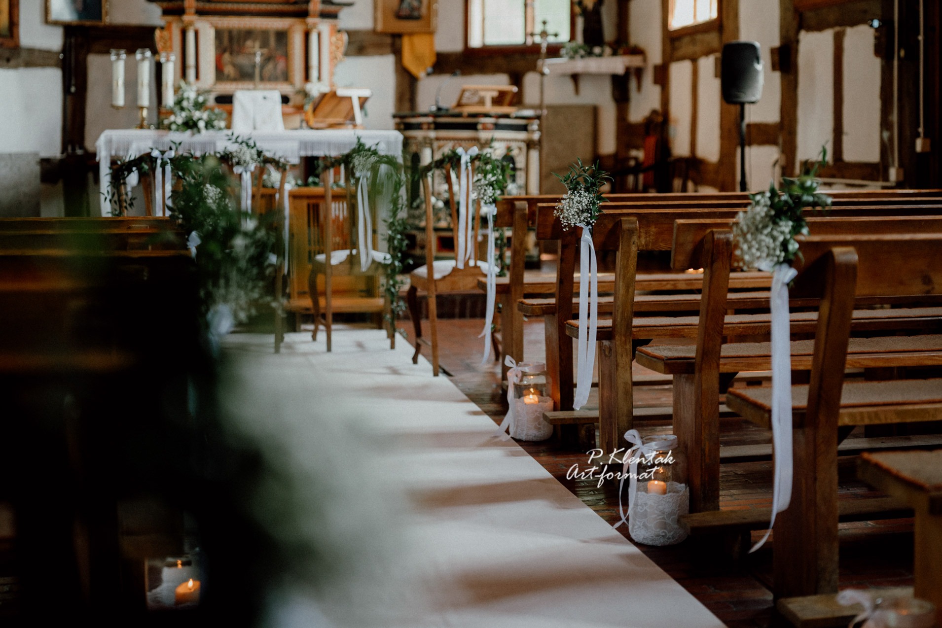 Udekorowany kościół przed ceremonią ślubną: biały dywan, drewniane ławki z kwiatami i wstążkami, lampiony ze świecami, ołtarz w tle.