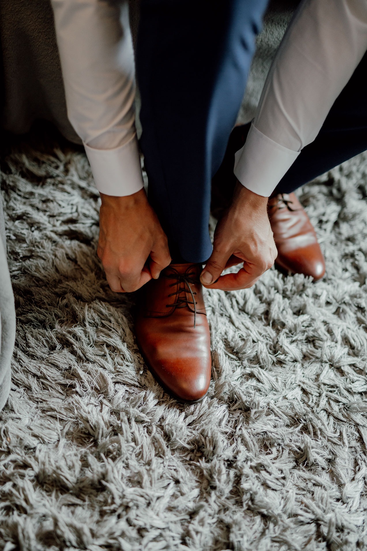 Mężczyzna w granatowym garniturze wiąże sznurowadła brązowych skórzanych butów na puszystym, szarym dywanie.