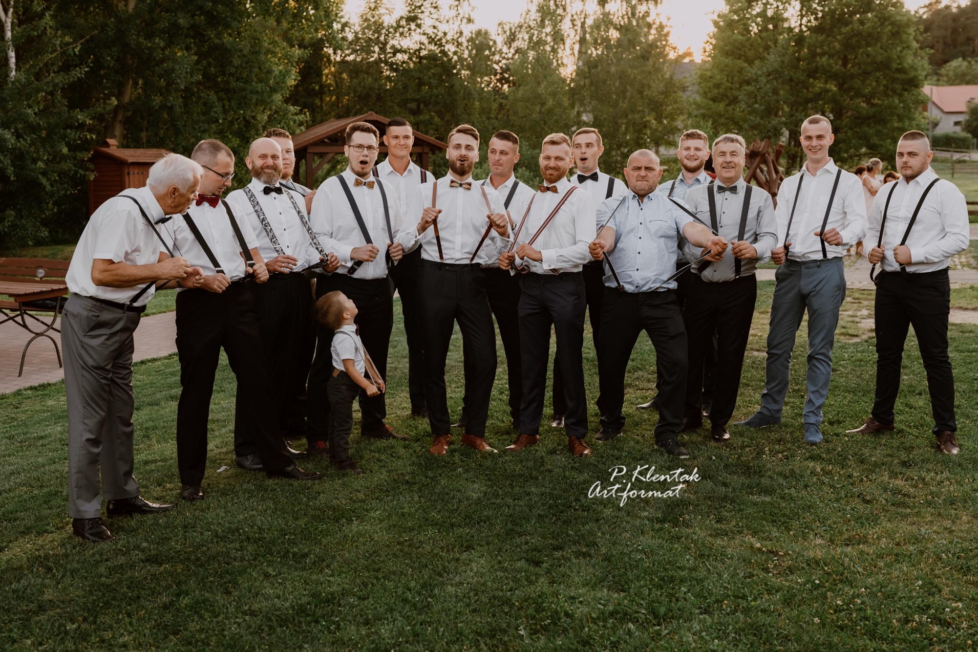 Grupa mężczyzn, w tym jeden mały chłopiec, pozuje do zdjęcia na zewnątrz, ubrani w białe koszule, czarne spodnie i szelki, z różnymi muszkami. Jeden z mężczyzn ma rozpiętą koszulę.