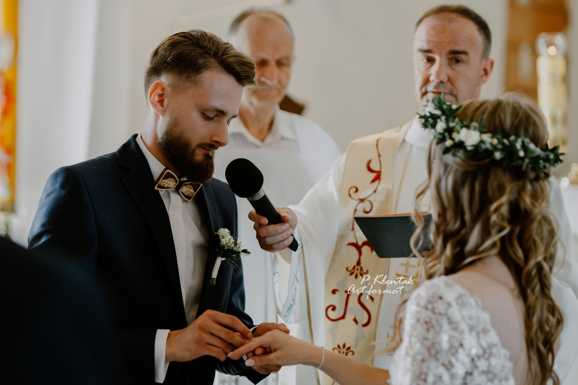 Moment zakładania obrączki podczas ceremonii ślubnej, pan młody w granatowym garniturze z drewnianą muszką, ksiądz w ornacie, panna młoda z wiankiem na głowie.
