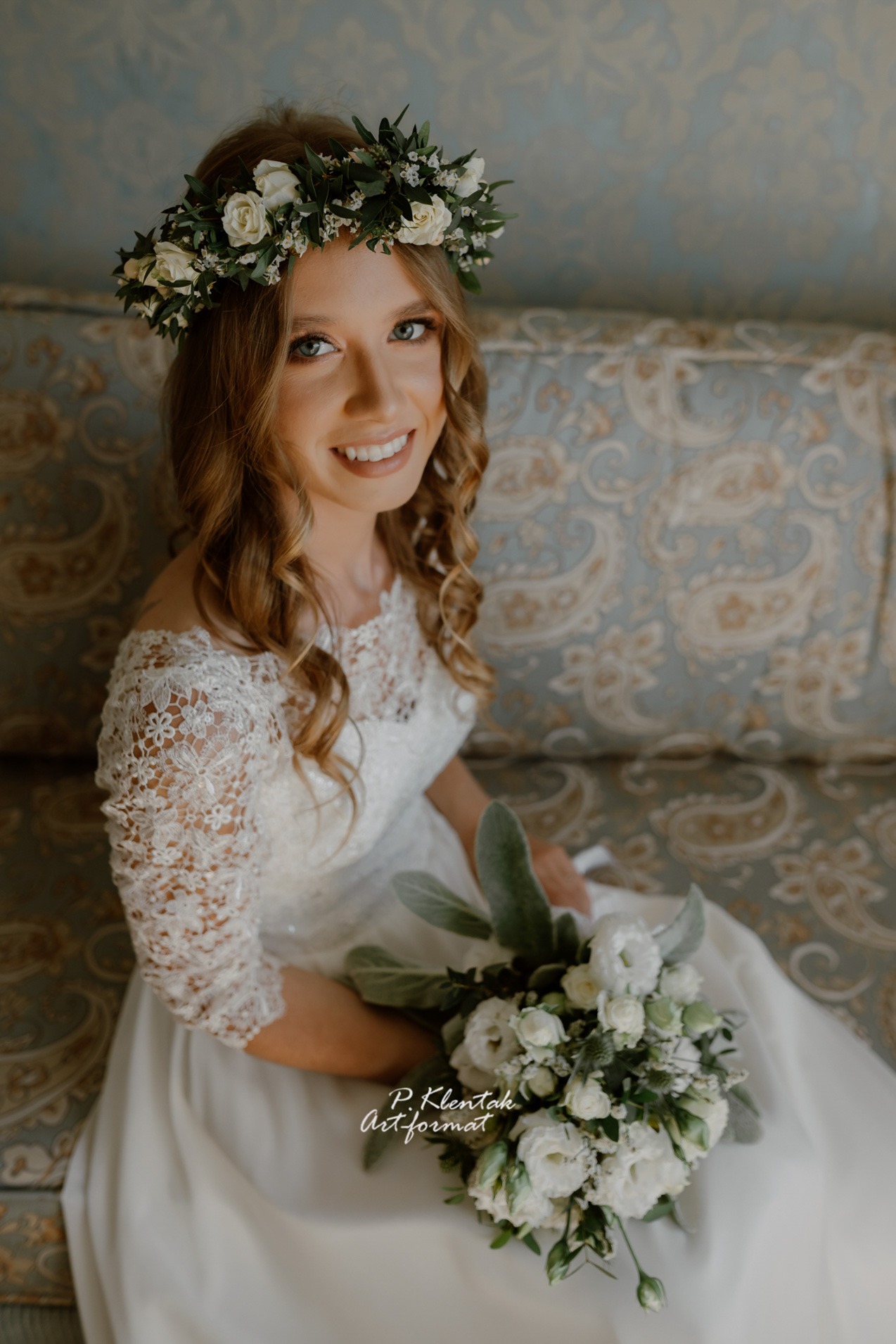 Uśmiechnięta panna młoda w koronie z białych róż i sukni z koronkowymi rękawami, siedząca na wzorzystej sofie i trzymająca bukiet z białych kwiatów.