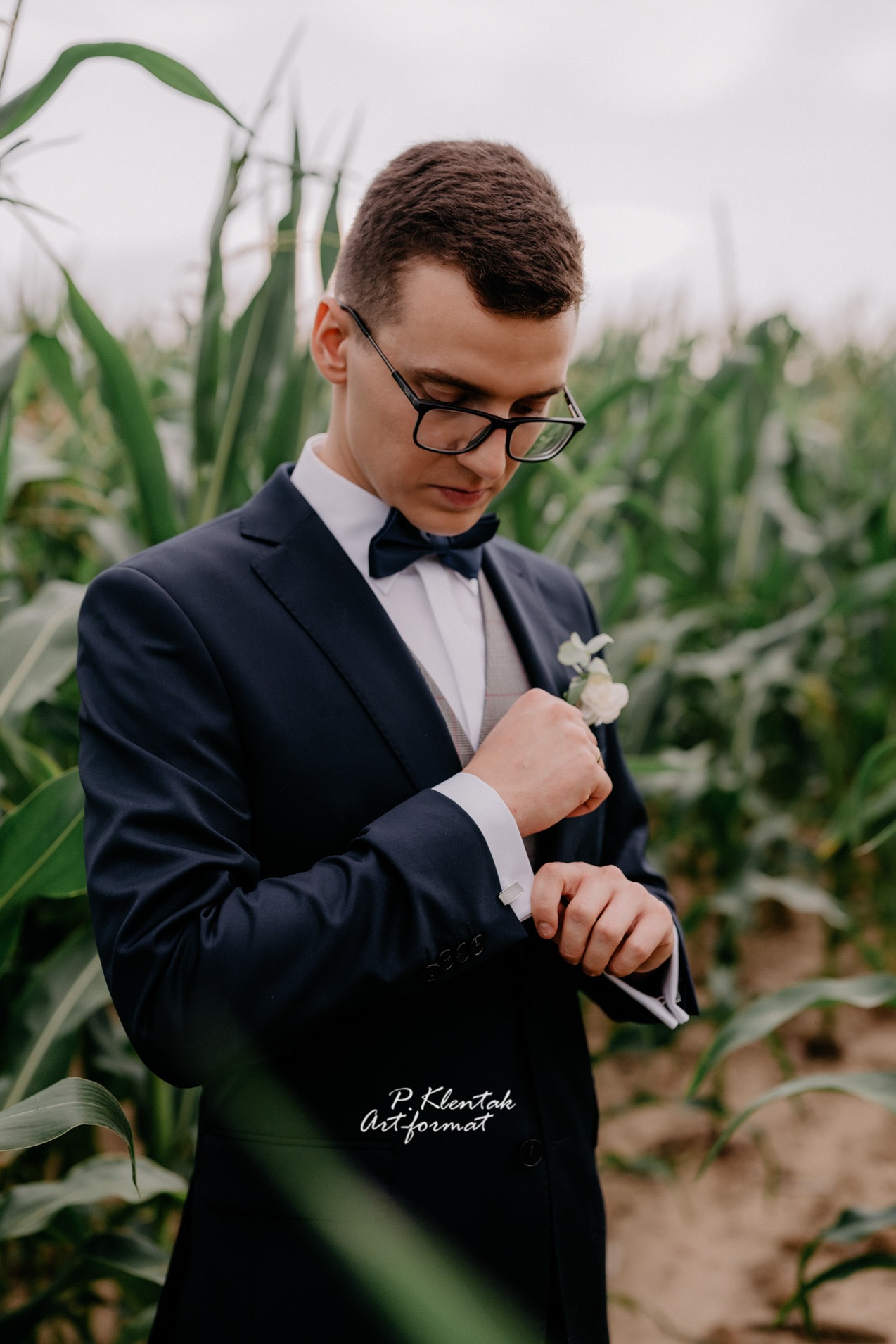 Młody mężczyzna w granatowym garniturze i muszce, poprawiający mankiet koszuli na tle pola kukurydzy, z delikatną butonierką w klapie.