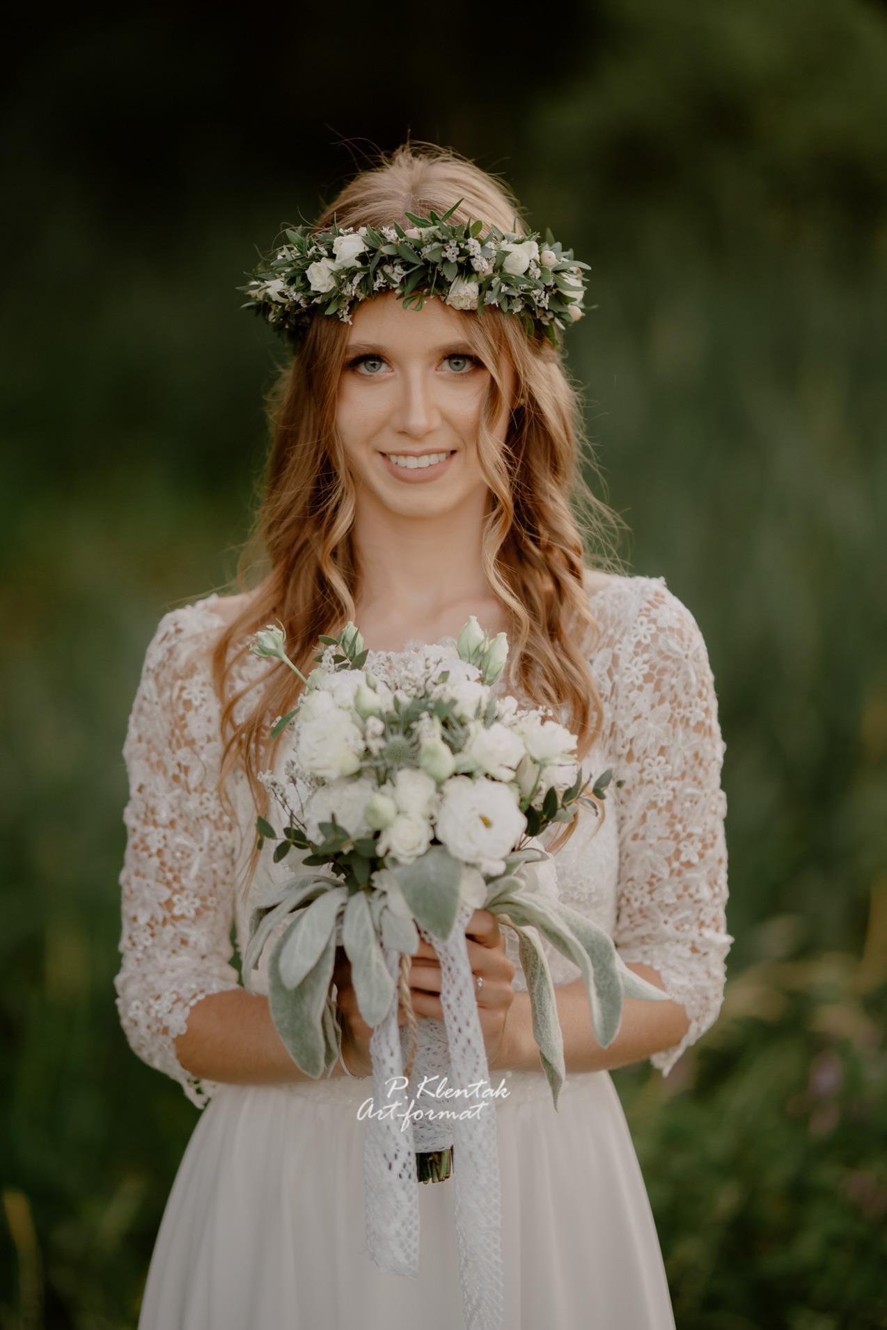 Uśmiechnięta panna młoda w koronie z białych kwiatów i sukni z koronkowymi rękawami, trzyma bukiet ślubny z białych kwiatów i zielonych liści z ozdobną wstążką.