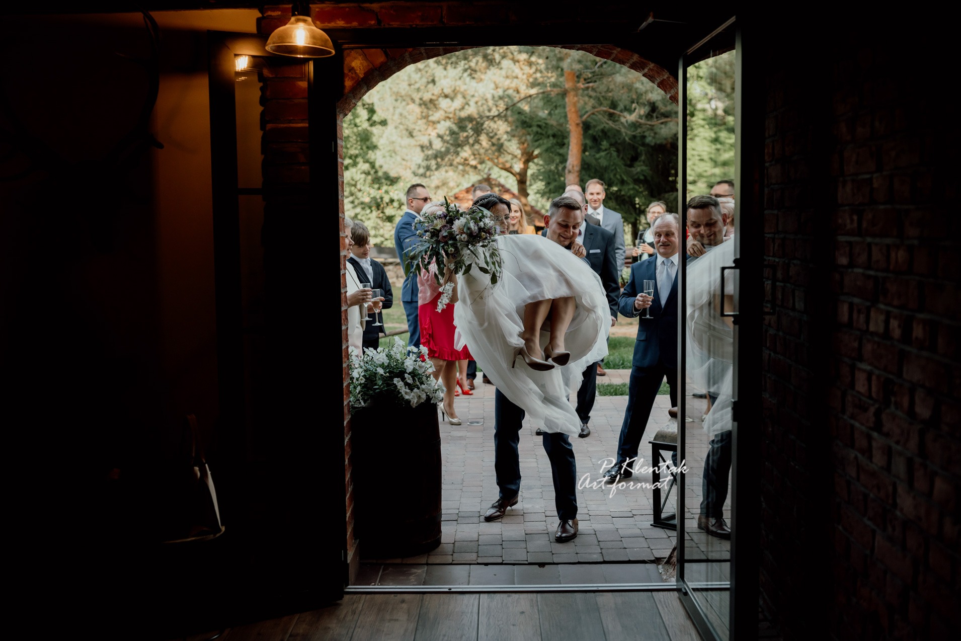 Pan młody niesie pannę młodą przez próg, trzymając ją na rękach, z bukietem kwiatów w dłoni, goście weselni z kieliszkami szampana w tle, widoczne brązowe skórzane buty pana młodego.