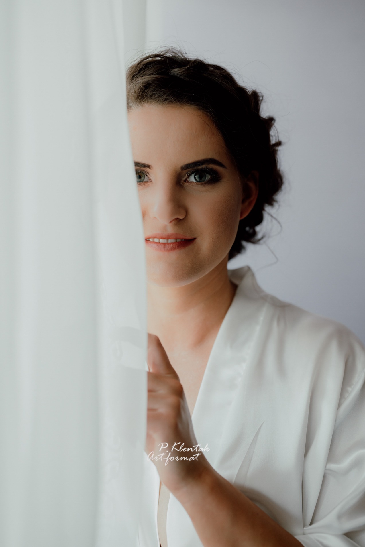 Portret panny młodej w białym szlafroku, częściowo zasłoniętej zwiewną firanką, z delikatnym makijażem i upiętymi włosami, patrzącej w obiektyw.