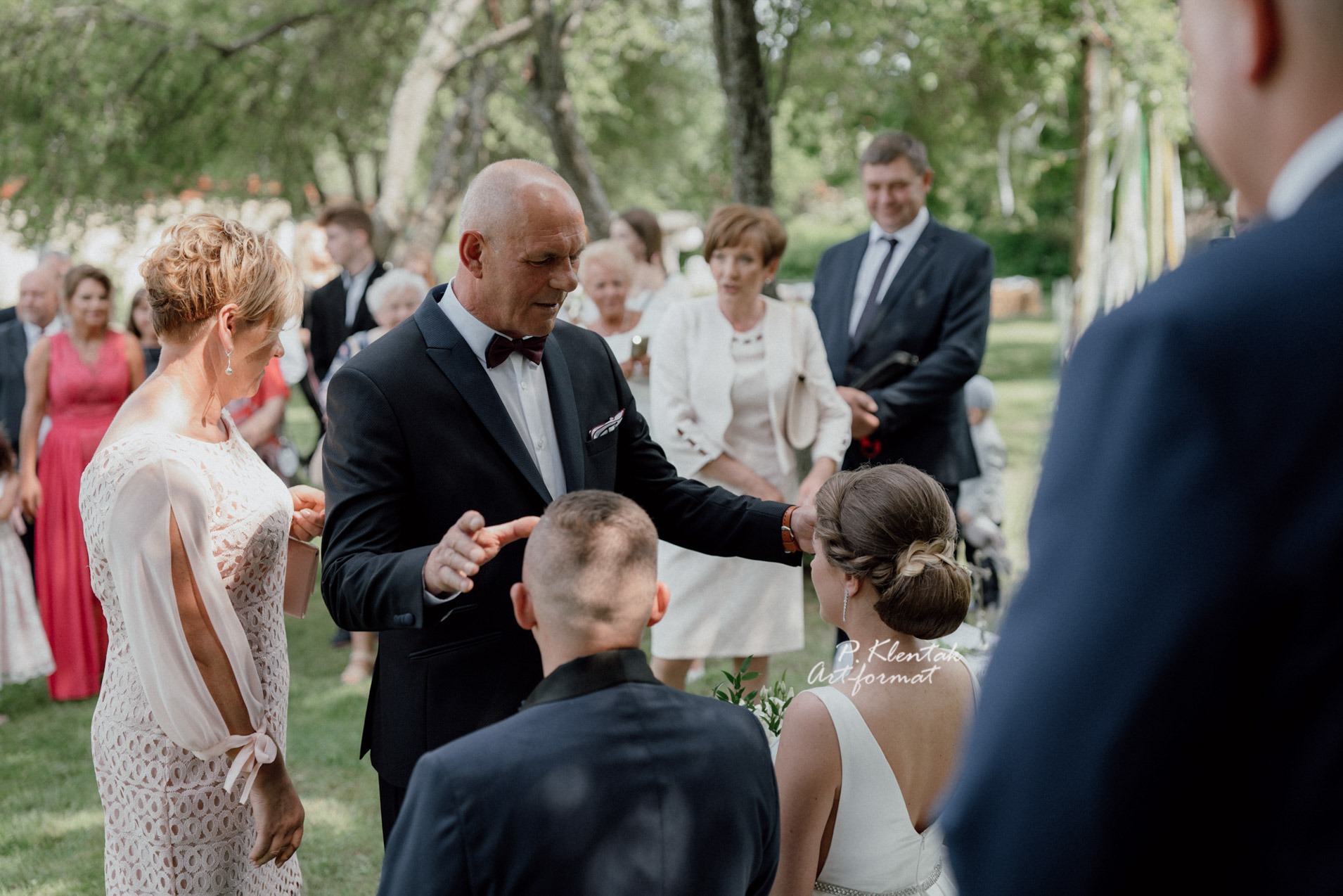 Uroczystość ślubna w plenerze: mężczyzna w garniturze z muszką błogosławi parę młodą, w tle goście i drzewa.