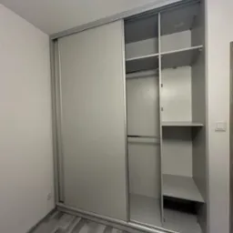 Jasnoszara szafa wnękowa z przesuwnymi drzwiami, widoczne p&oacute;łki i drążek na ubrania, podłoga w odcieniach szarości imitująca drewno.