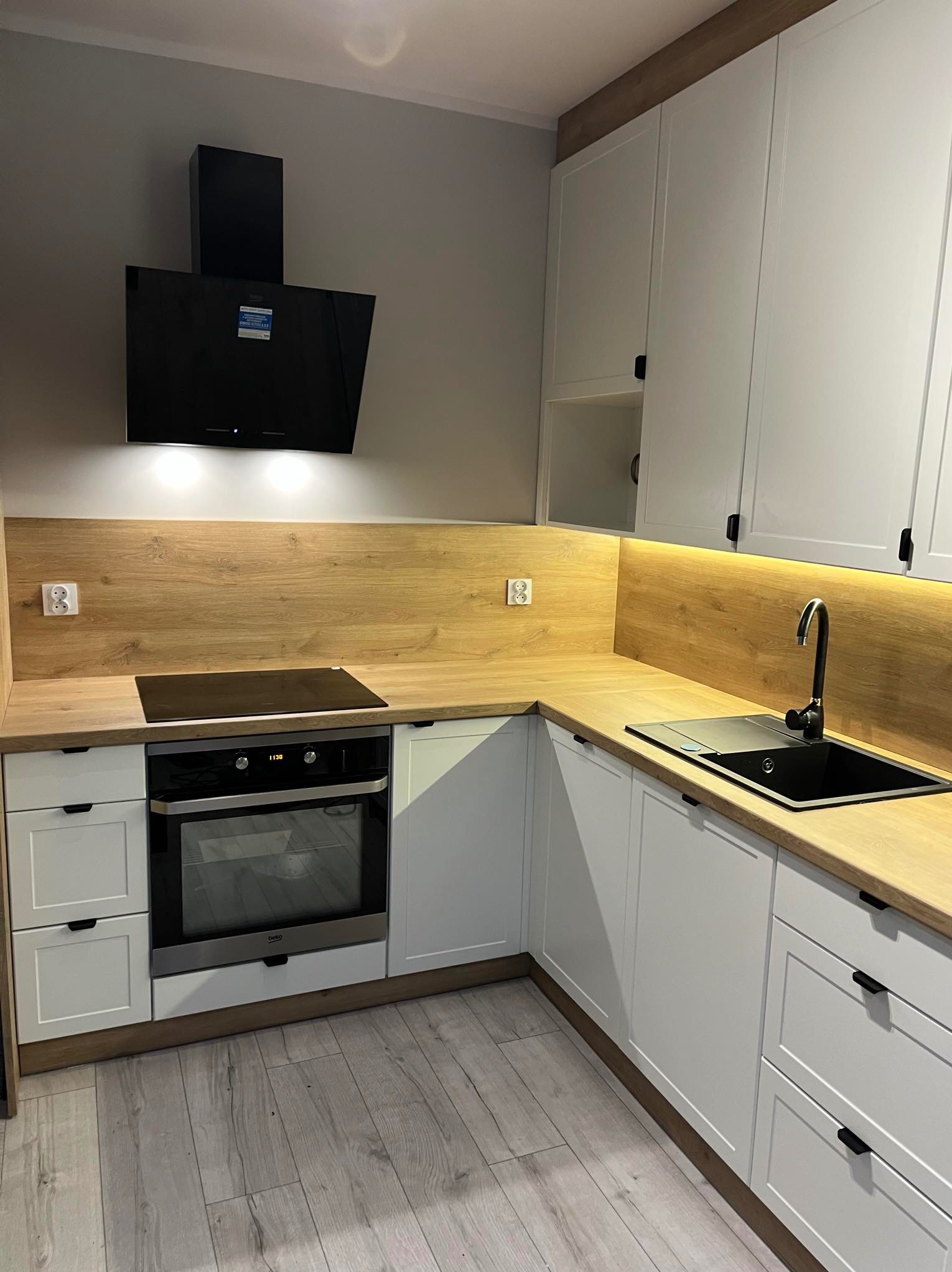 Nowoczesna kuchnia z białymi szafkami, drewnianym blatem, czarnym zlewem i okapem, oświetleniem LED pod szafkami wiszącymi i płytą indukcyjną nad piekarnikiem