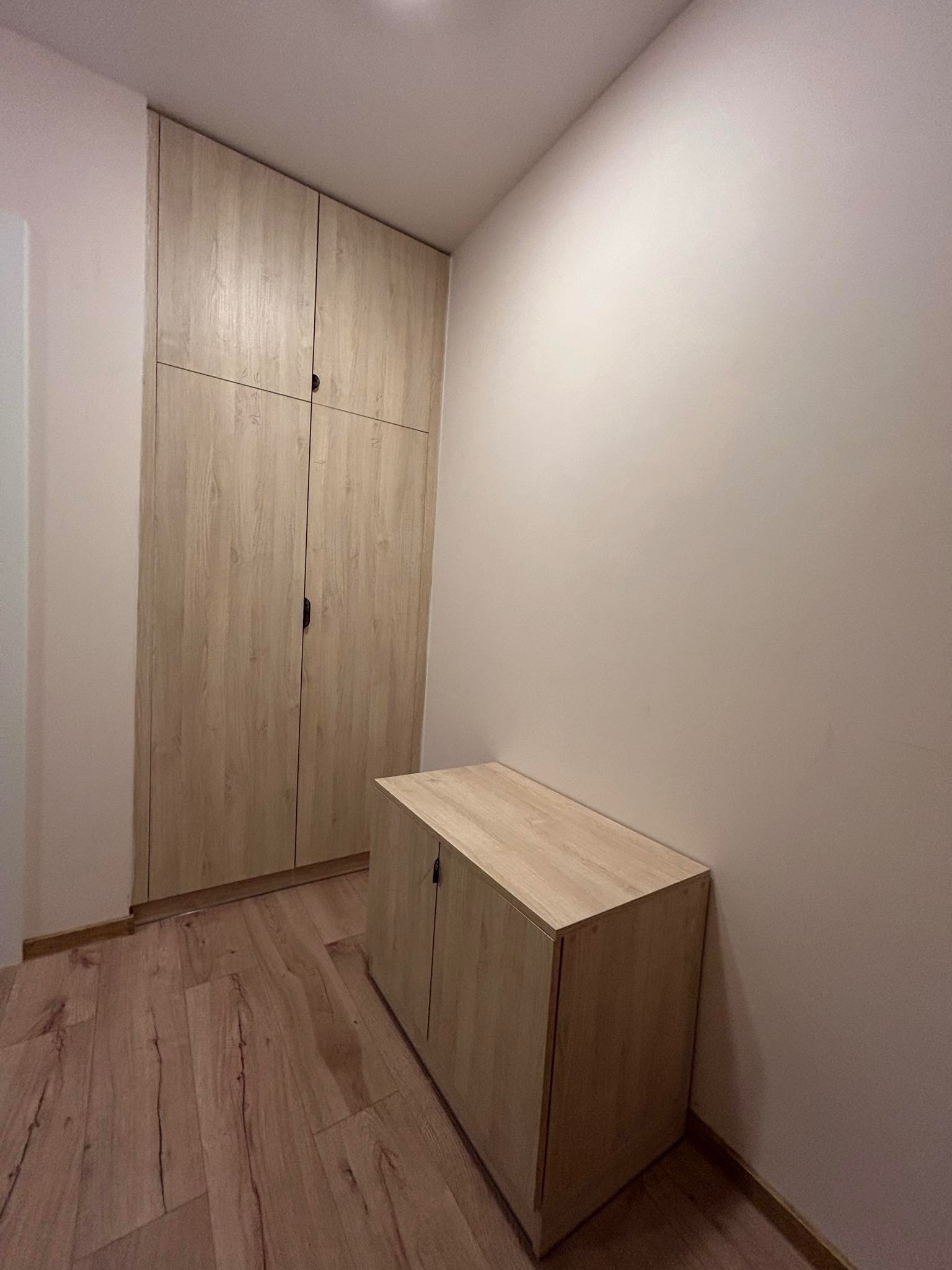 Wysoka szafa wnękowa z drewnopodobnymi frontami, dopasowana do wysokości pomieszczenia, oraz niska szafka zamykana na klucz, na drewnianej podłodze w jasnym korytarzu.
