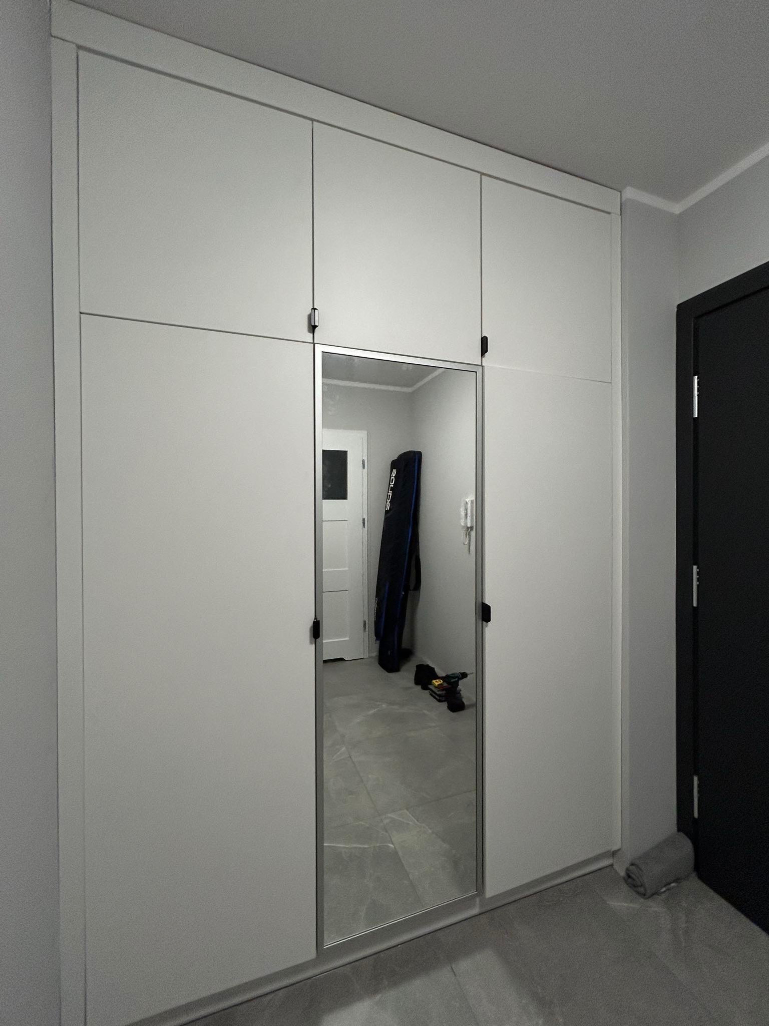 Biała szafa wnękowa z lustrzanymi drzwiami w przedpokoju, widoczny wiertarko-wkrętak i pokrowiec na sprzęt sportowy odbijające się w lustrze.