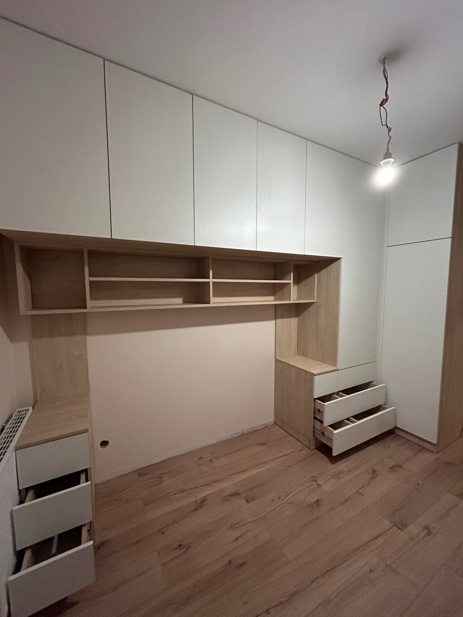 Biało-drewniana zabudowa meblowa w pokoju z otwartymi szufladami i półkami, widoczne drewniane panele podłogowe i wisząca żarówka.