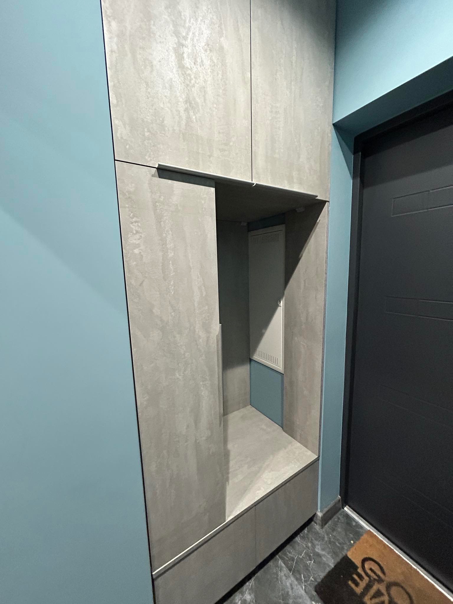 Wnęka z szarą szafką z otwartą półką i siedziskiem, obok ciemnych drzwi wejściowych i turkusowej ściany.