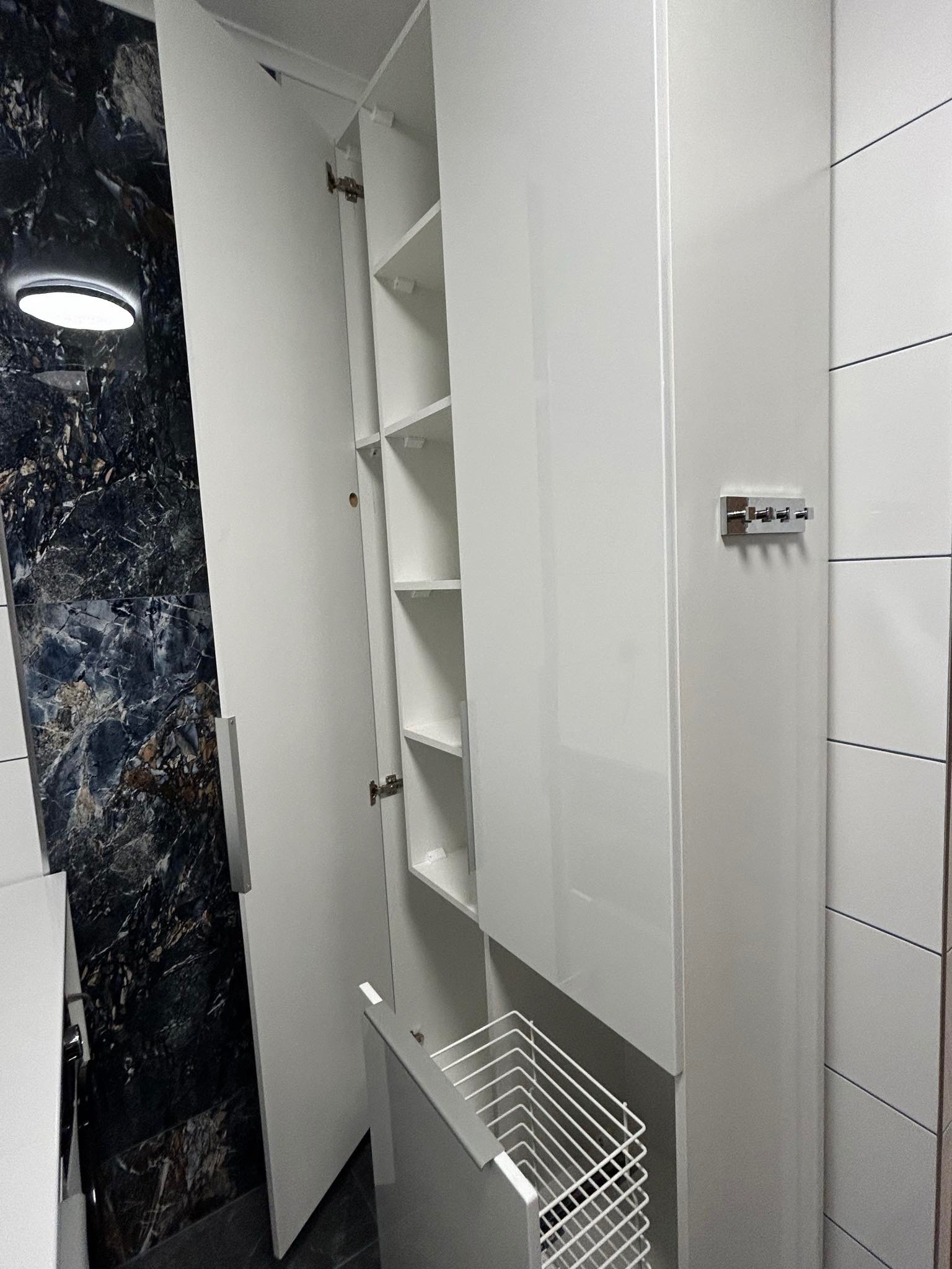 Biała, lakierowana szafa wnękowa z otwartymi drzwiami, ukazująca półki i wysuwany kosz na pranie, na tle ściany z czarnego marmuru i płytek.