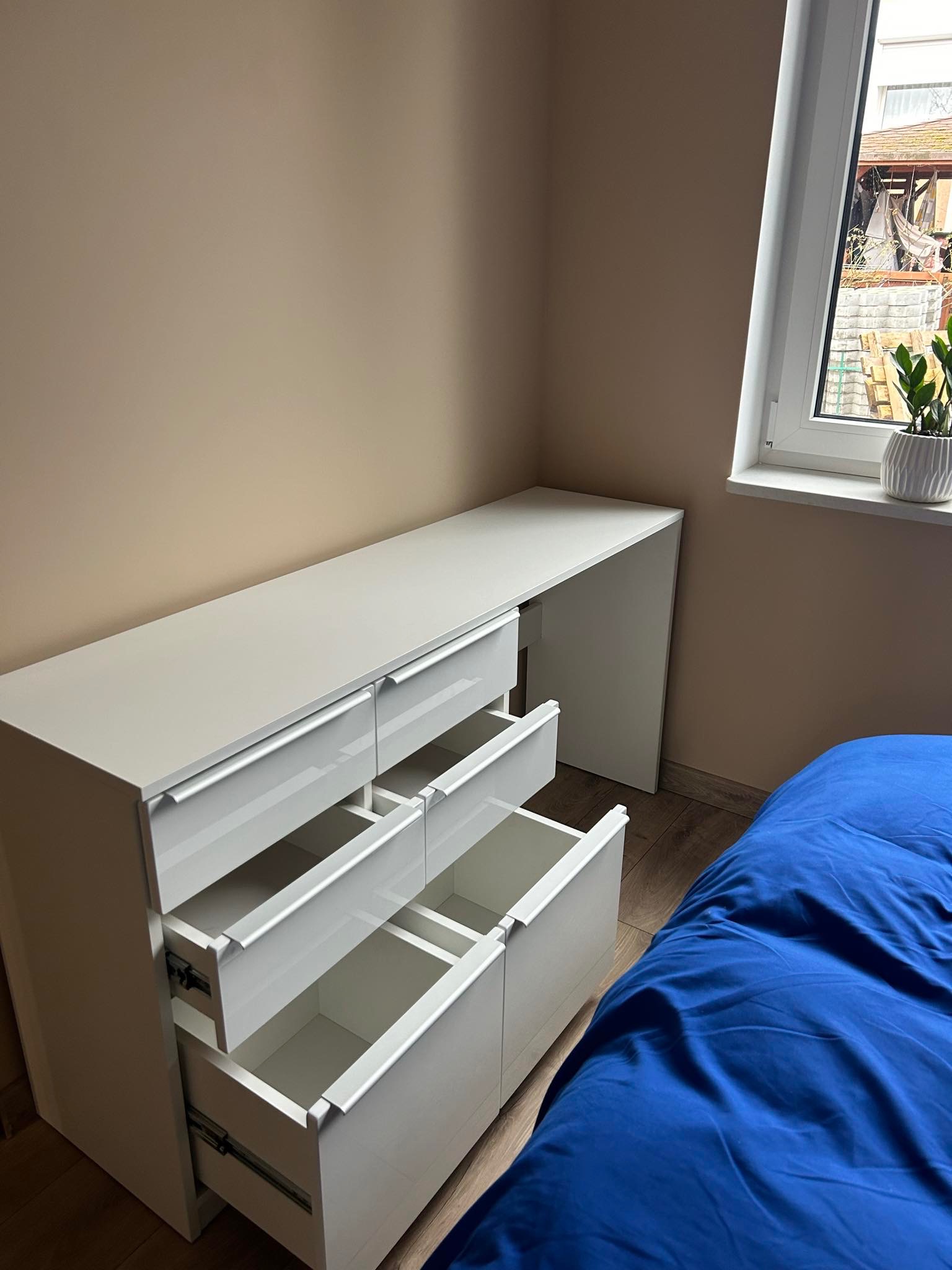 Białe biurko z szufladami na tle beżowej ściany, trzy szuflady otwarte, obok niebieska pościel, widok na okno z rośliną.