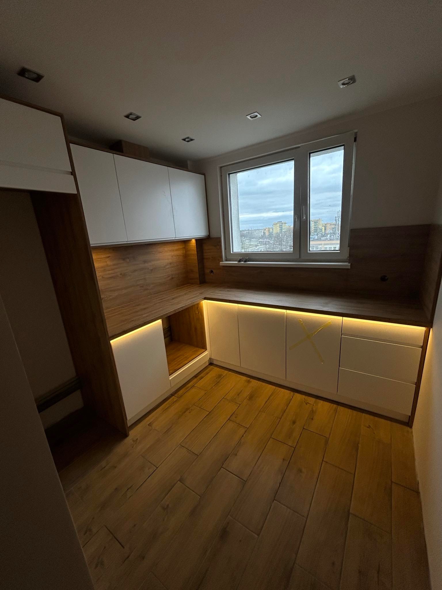 Nowoczesna kuchnia z białymi szafkami i drewnianymi akcentami, oświetleniem LED pod szafkami wiszącymi i blatem, widok na miasto przez okno, zabezpieczenia transportowe na frontach szafek.