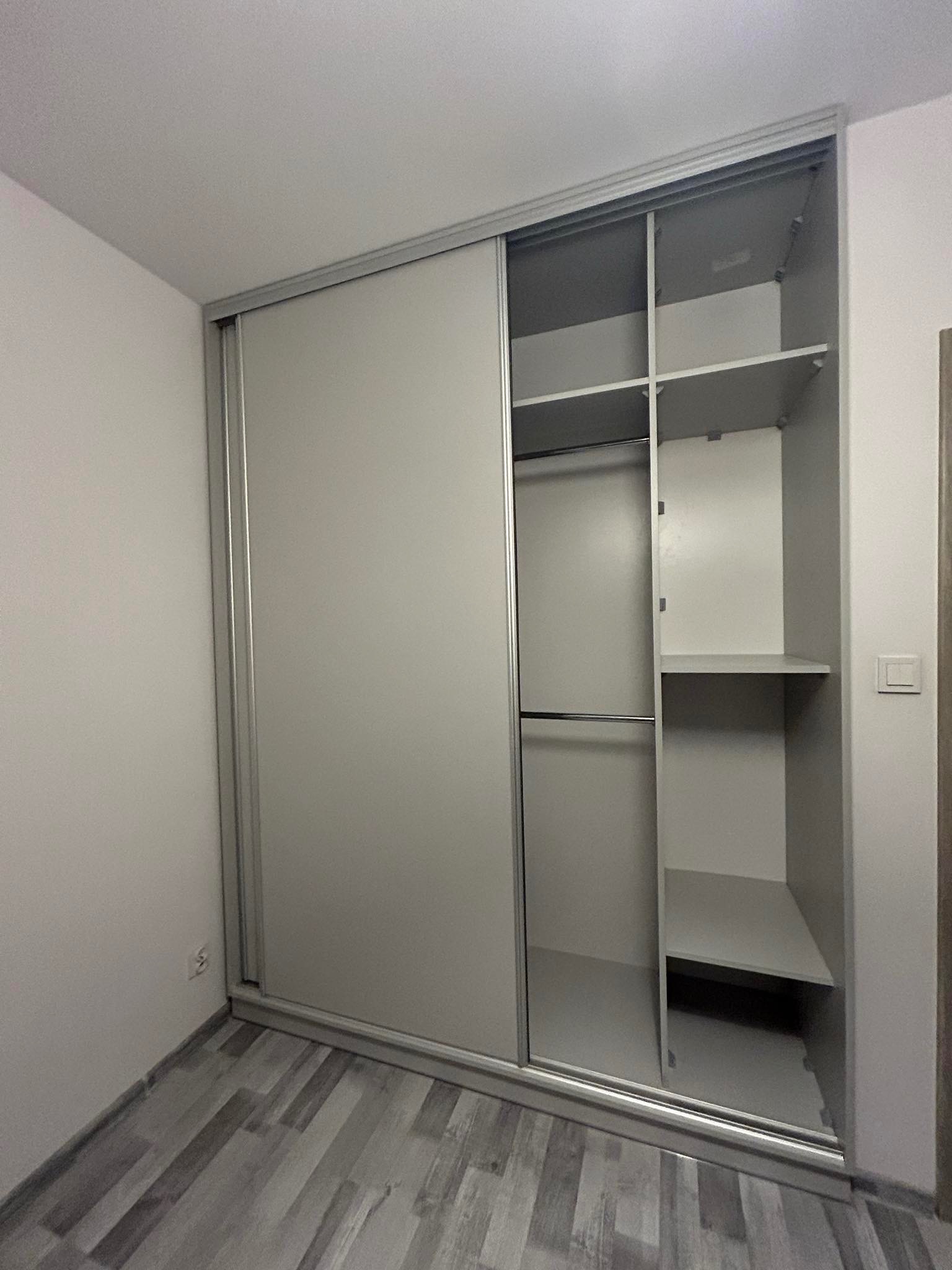 Jasnoszara szafa wnękowa z przesuwnymi drzwiami, widoczne półki i drążek na ubrania, podłoga w odcieniach szarości imitująca drewno.