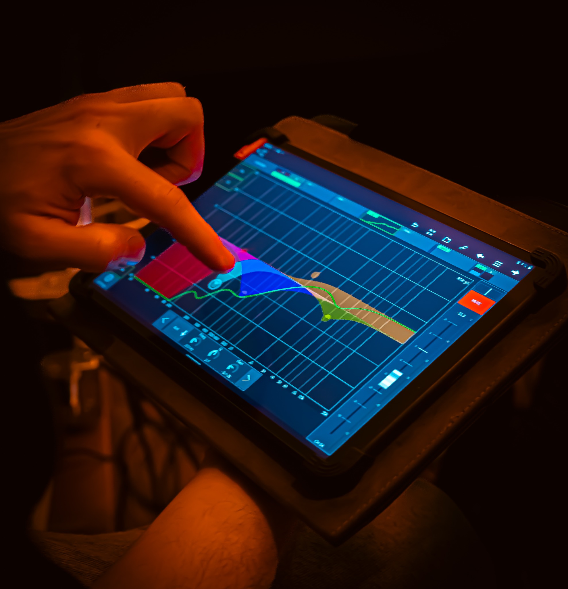 Palec dotyka ekranu tabletu z aplikacją do miksowania dźwięku, wyświetlającą kolorowy equalizer z siatką, w scenerii przyciemnionego otoczenia.
