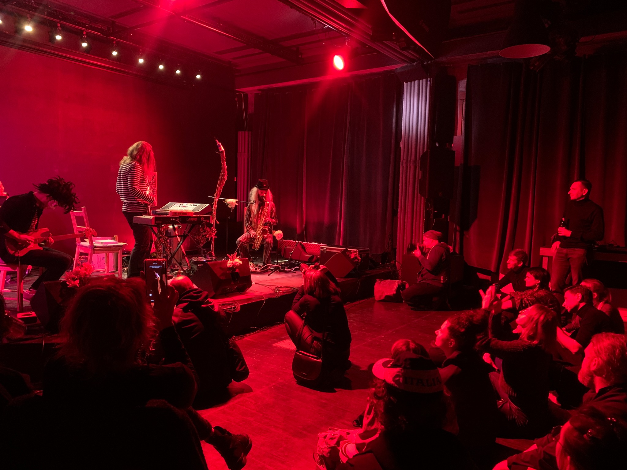 Zespół muzyczny grający na scenie oświetlonej czerwonym światłem, widownia siedzi na podłodze, jeden z muzyków gra na saksofonie z ozdobami.