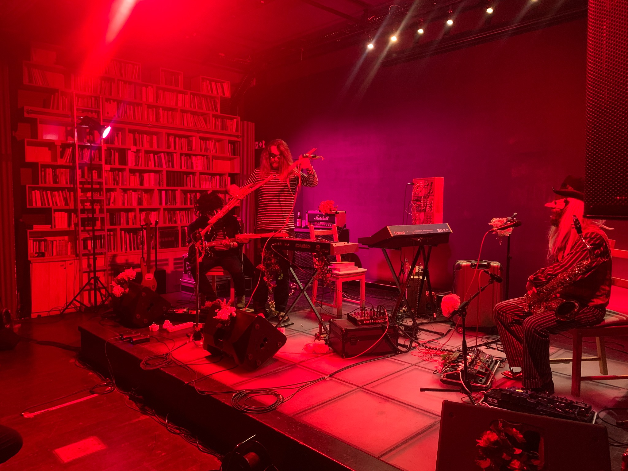 Zespół muzyczny podczas występu na scenie w czerwonym oświetleniu, widoczny keyboard, gitara, saksofon, kolumny głośnikowe i wzmacniacze, w tle ściana z książkami.