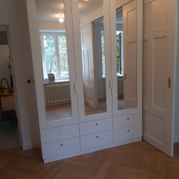 ATUMEBEL - Biała szafa wnękowa z lustrami na frontach, widoczne złote gałki szuflad, parkiet w jodełkę na podłodze, odbicie wnętrza pokoju w lustrach.