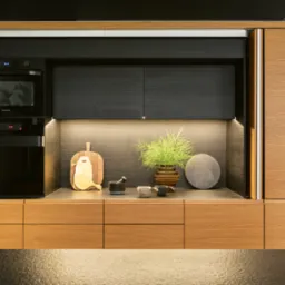 Nowoczesna zabudowa kuchenna z drewnianymi frontami w ciepłym odcieniu, czarnymi elementami i wbudowanym piekarnikiem. Na blacie dekoracje: deski do krojenia, zioła w doniczce i kamienne podkładki.