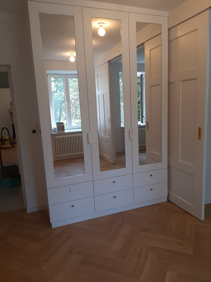 Biała szafa wnękowa z lustrami na frontach, widoczne złote gałki szuflad, parkiet w jodełkę na podłodze, odbicie wnętrza pokoju w lustrach.