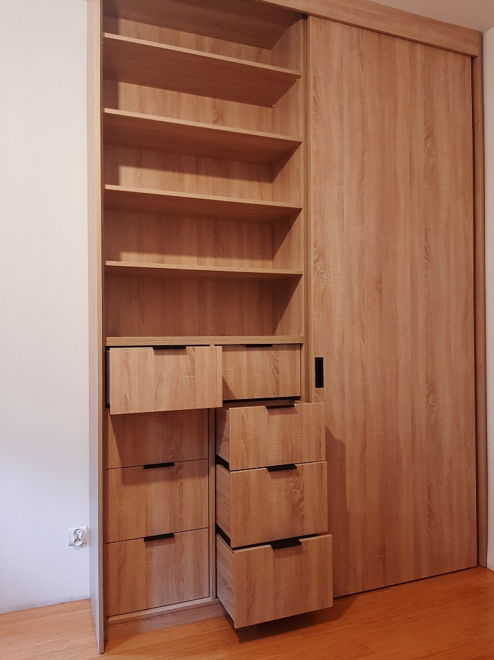 Jasna, drewnopodobna szafa wnękowa z otwartymi półkami i wysuwanymi szufladami z czarnymi uchwytami, zamontowana w mieszkaniu w Warszawie.