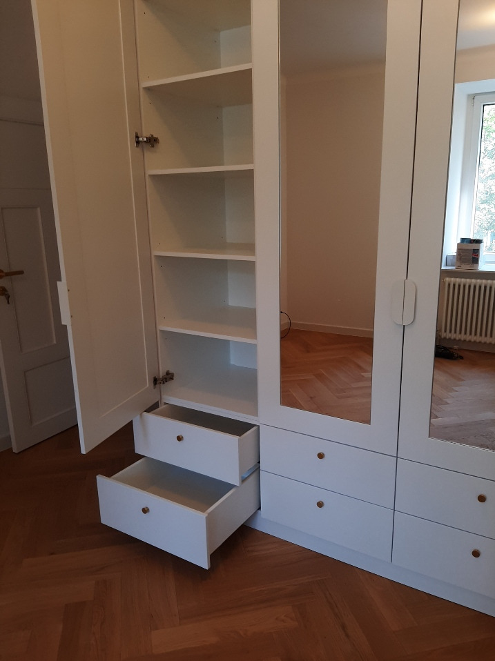 Biała szafa z otwartymi drzwiami i wysuniętymi szufladami, widoczne półki i lustrzane fronty, na podłodze parkiet w jodełkę.