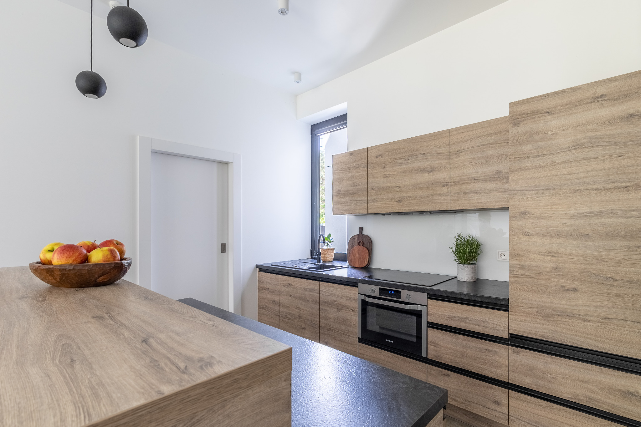 Nowoczesna kuchnia z drewnianymi frontami szafek, czarnym blatem i oświetleniem punktowym. Na wyspie drewniana misa z jabłkami.