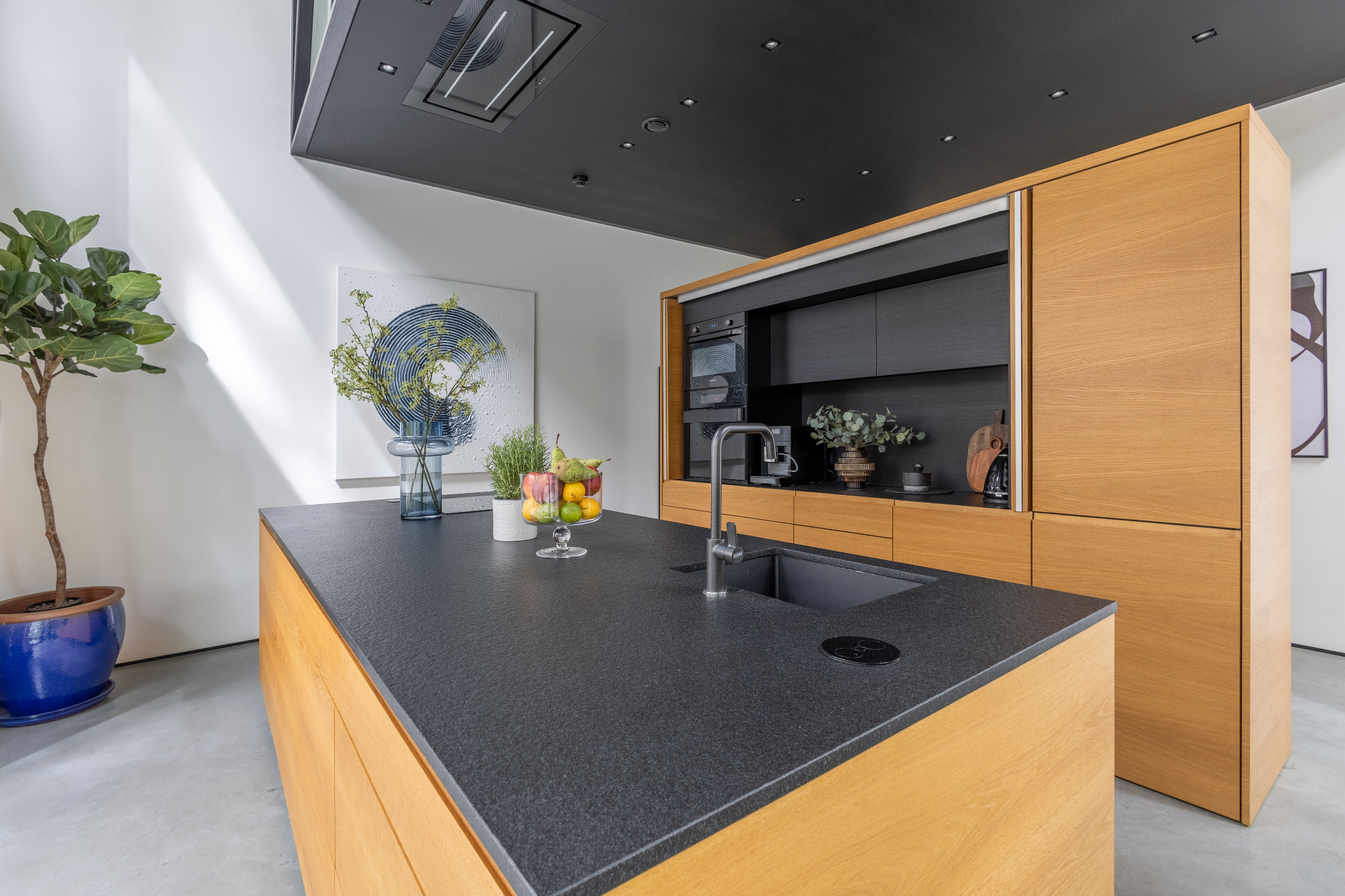 Nowoczesna kuchnia z wyspą i czarnym blatem z kamienia, drewniane fronty szafek, oświetlenie punktowe, dekoracje roślinne i owocowe.