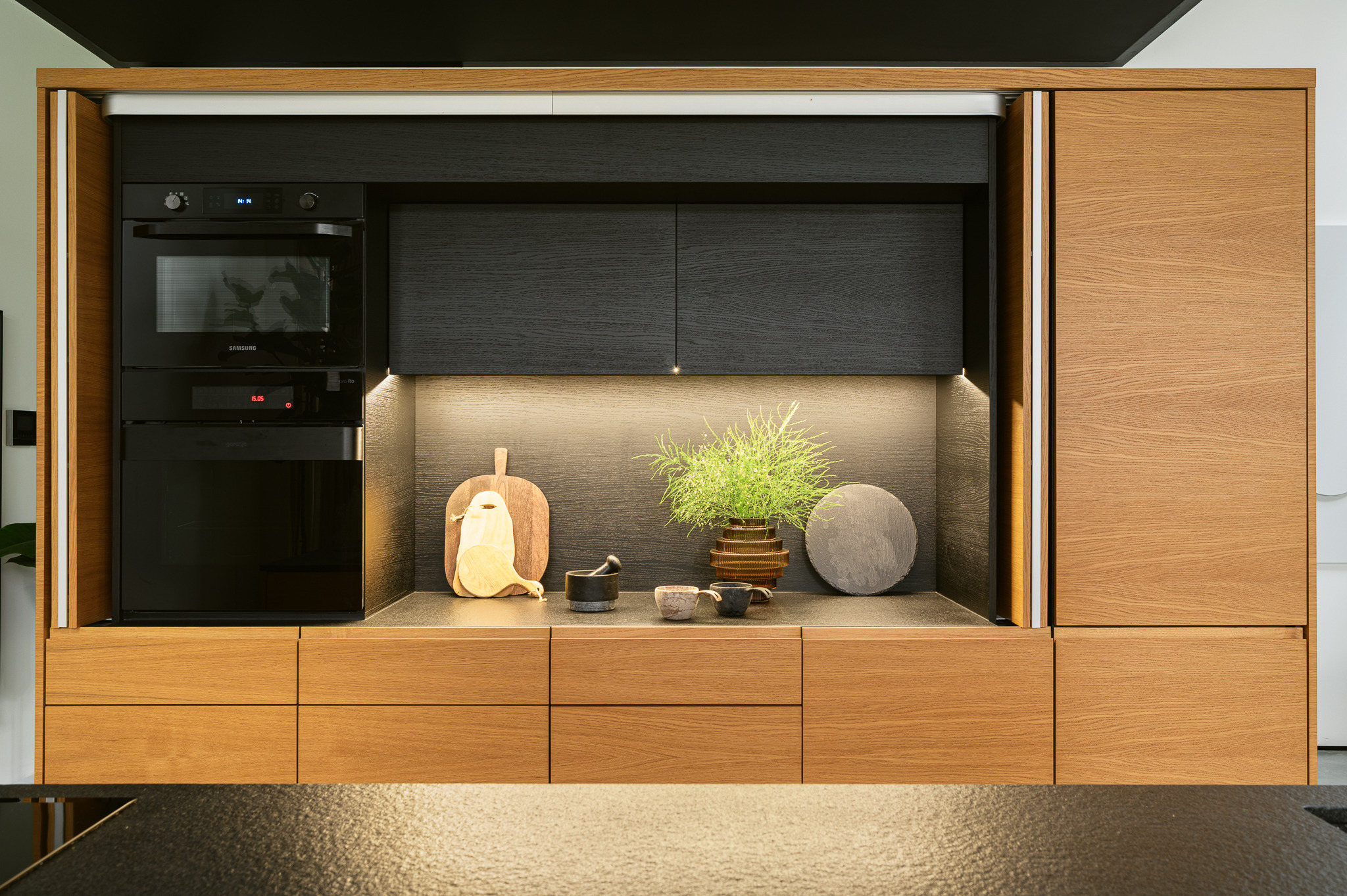 Nowoczesna zabudowa kuchenna z drewnianymi frontami w ciepłym odcieniu, czarnymi elementami i wbudowanym piekarnikiem. Na blacie dekoracje: deski do krojenia, zioła w doniczce i kamienne podkładki.