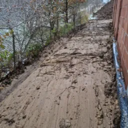 Udeptana, błotnista droga gruntowa po robotach ziemnych, z widocznymi śladami opon, prowadząca obok ściany budynku w trakcie budowy i zasypanej ziemią skarpy, w tle drzewa z resztkami śniegu.