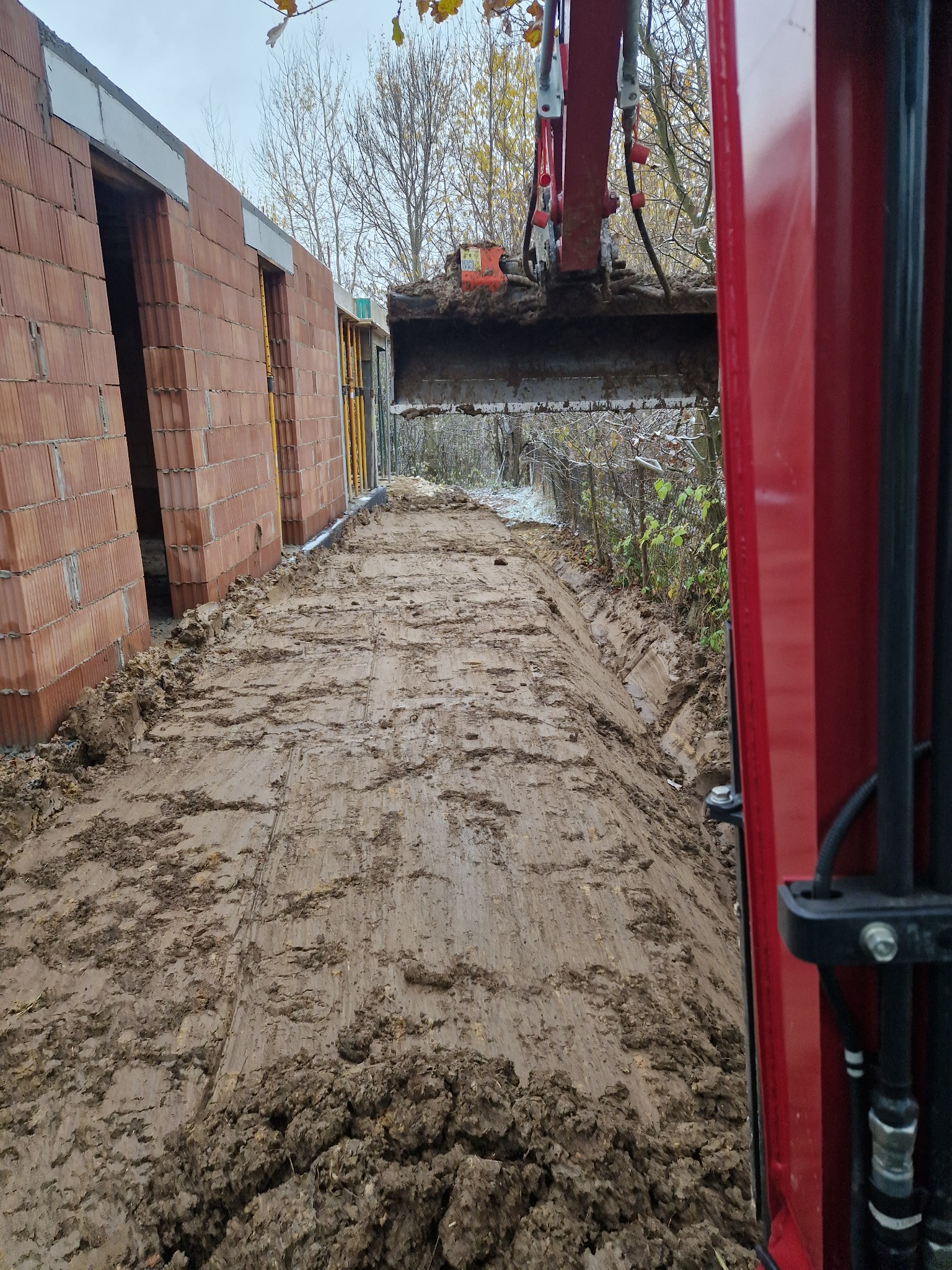 Czerwona koparka w trakcie prac ziemnych, widok z kabiny operatora na wykop wzdłuż fundamentów budynku w trakcie budowy, błotnisty teren.