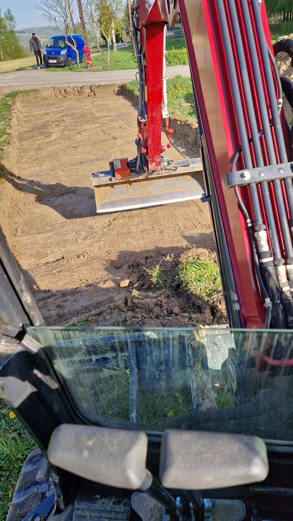 Widok z kabiny koparki na wykopany prostokątny dół w ziemi, z widoczną czerwoną konstrukcją ramienia koparki i osobą oraz niebieskim busem w tle.