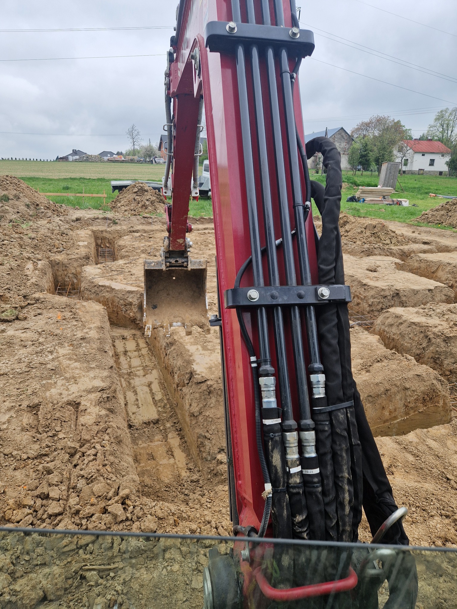 Widok z kabiny koparki na wykop pod fundamenty zbrojone siatką, w tle zabudowa wiejska i pochmurne niebo.