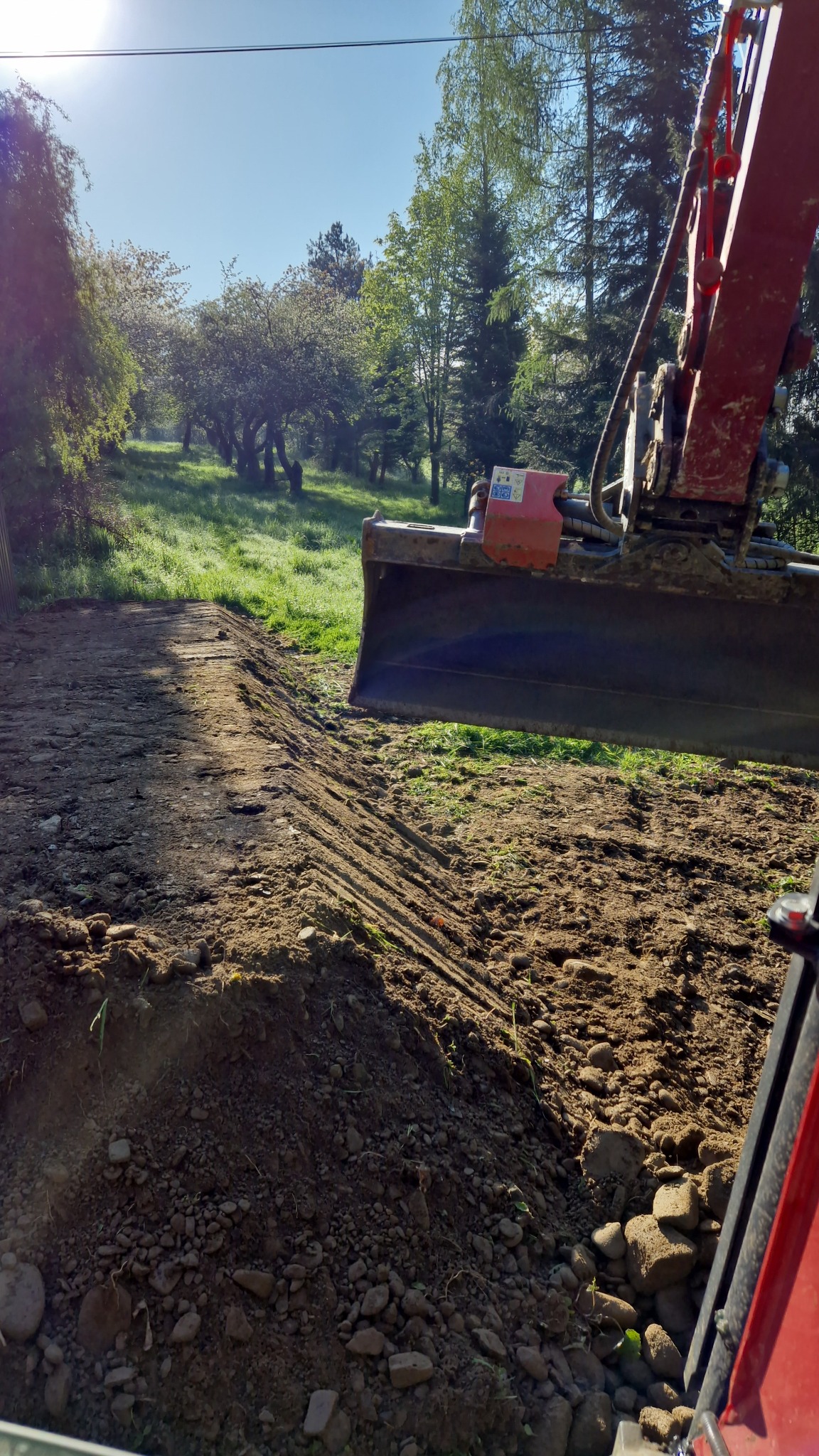 Czerwona koparka wyrównuje teren na tle zielonego wzgórza z drzewami.