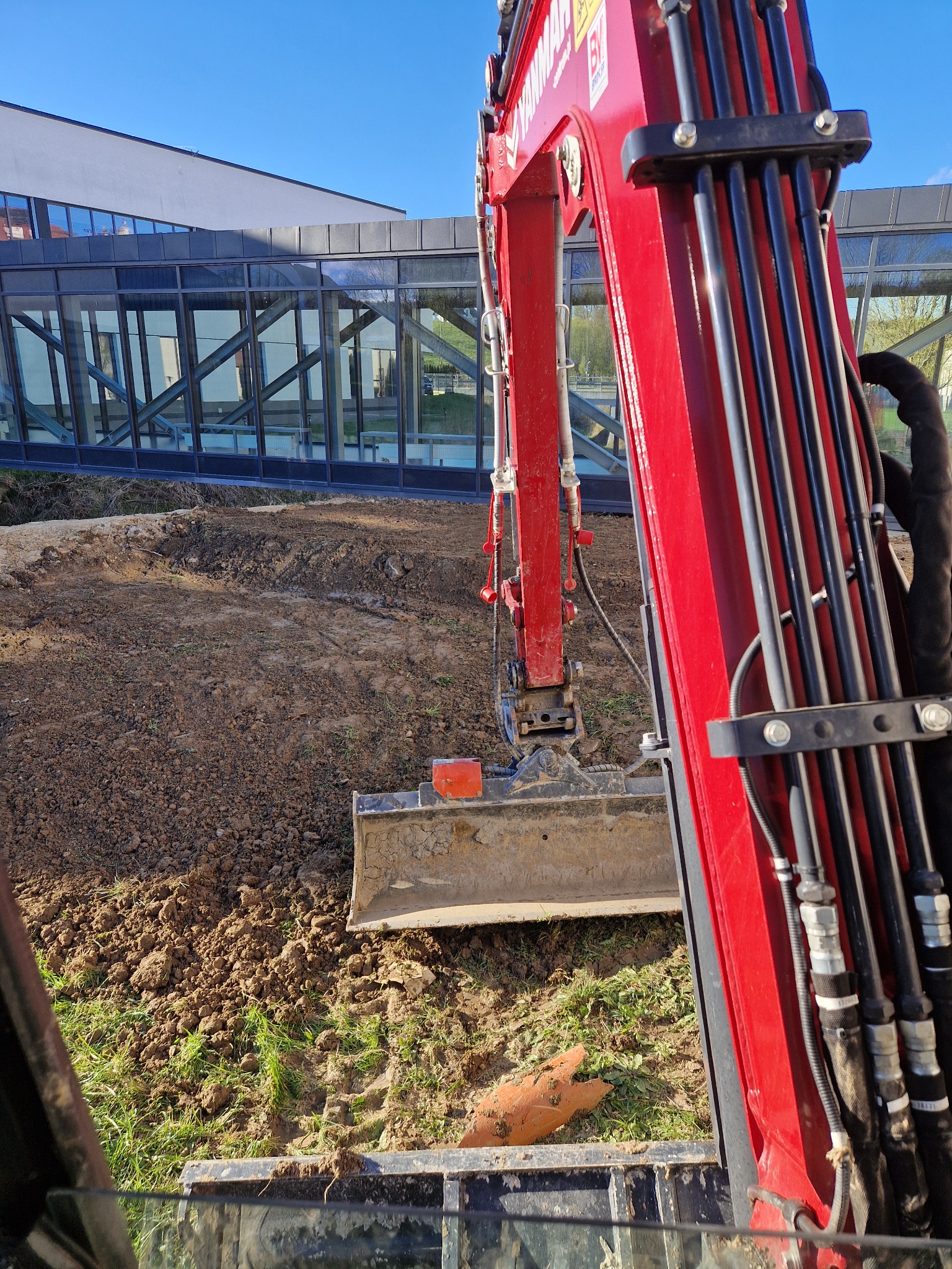 Czerwona koparka z łyżką w trakcie prac ziemnych, widok z kabiny, w tle nowoczesny budynek z przeszklonym łącznikiem.