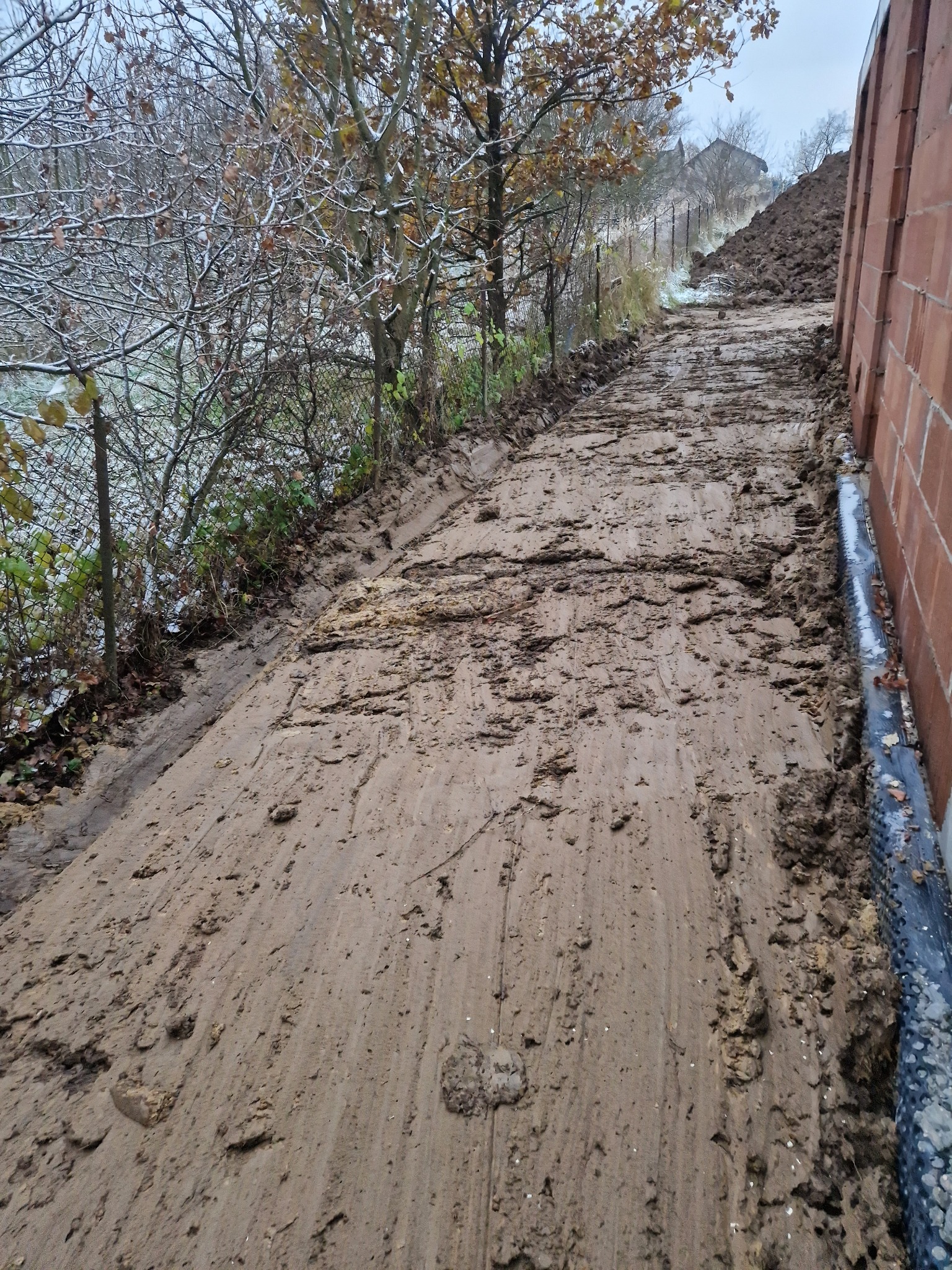 Udeptana, błotnista droga gruntowa po robotach ziemnych, z widocznymi śladami opon, prowadząca obok ściany budynku w trakcie budowy i zasypanej ziemią skarpy, w tle drzewa z resztkami śniegu.