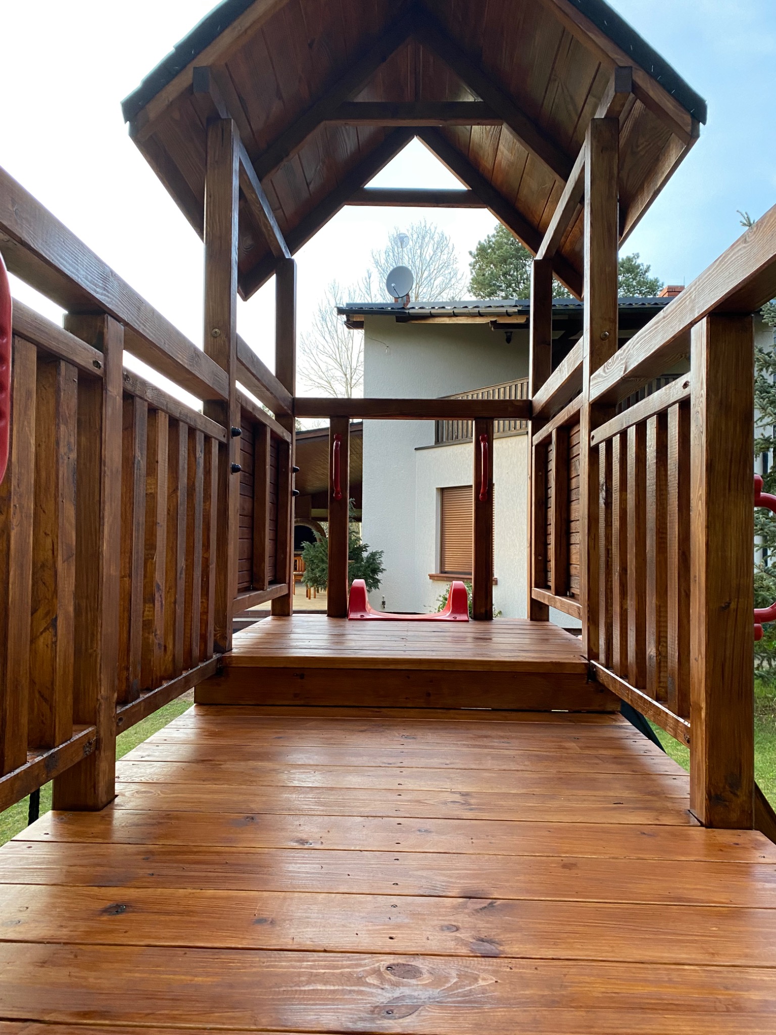 Widok z dołu na drewnianą konstrukcję placu zabaw z zadaszeniem, w tle dom z anteną satelitarną i zielenią.