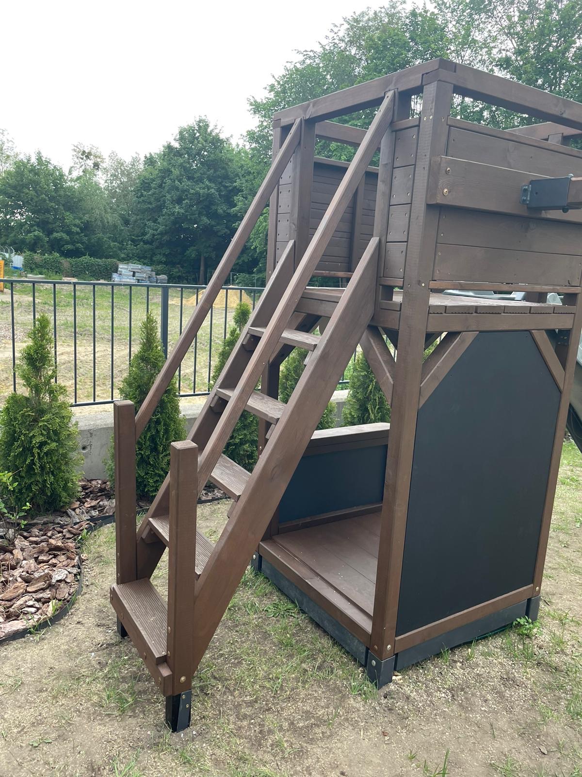 Brązowy drewniany domek dla dzieci z drabinką i tablicą do rysowania, otoczony tujami i korą drzewną.