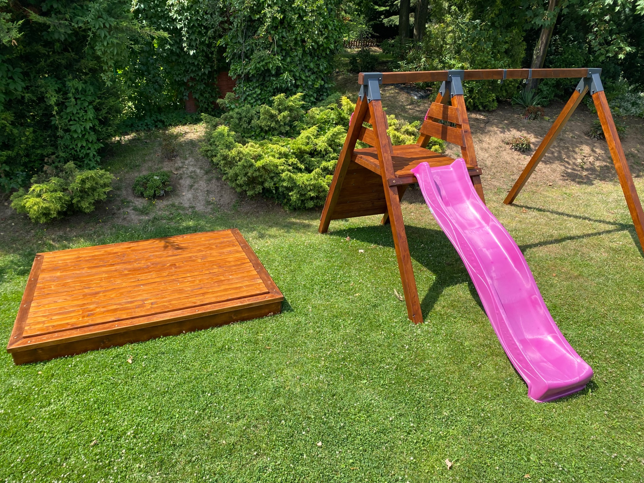Drewniany plac zabaw z piaskownicą i zjeżdżalnią w kolorze fuksji na zielonym trawniku, otoczony krzewami i drzewami.