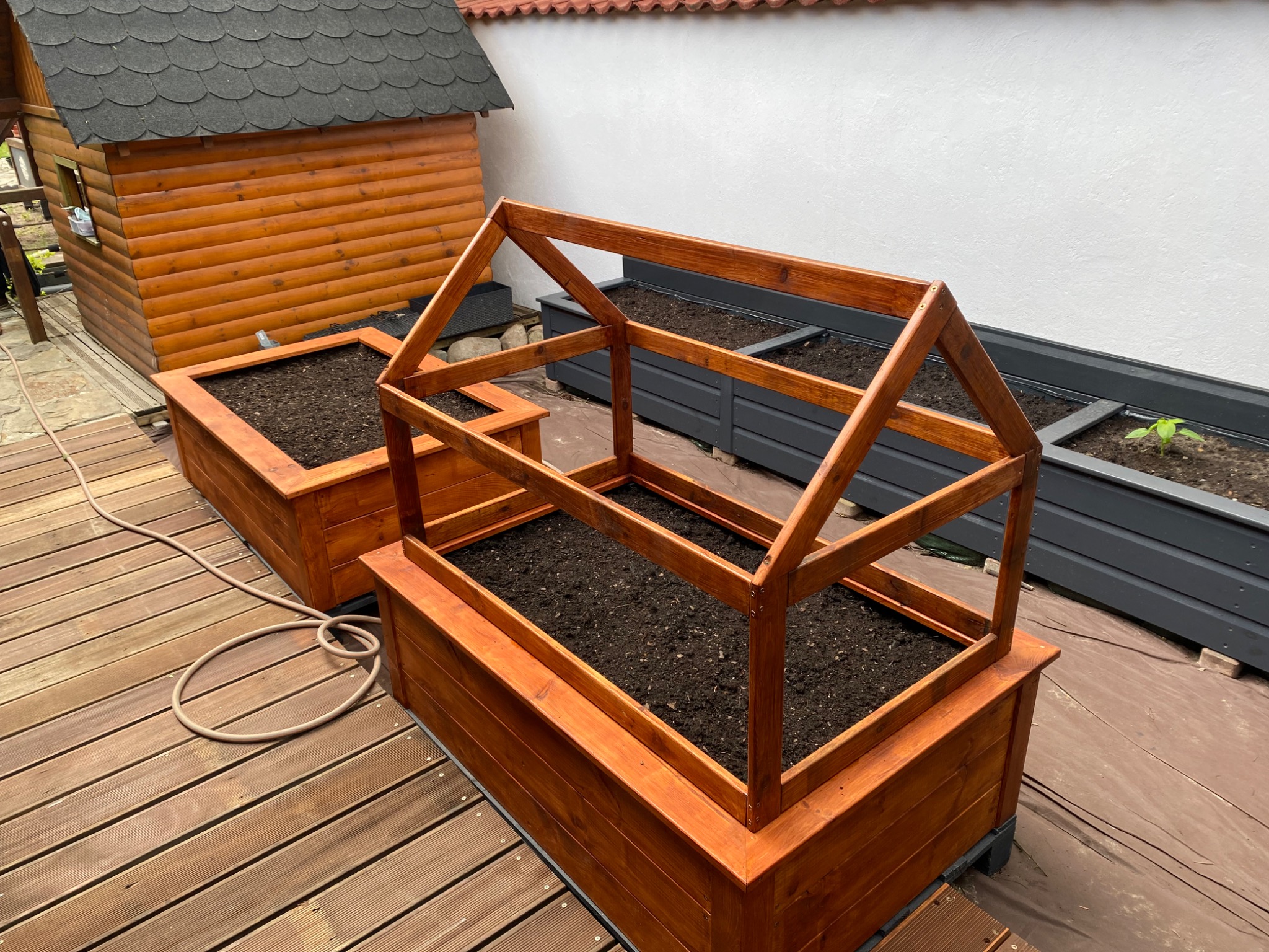 Drewniana konstrukcja szklarni na podwyższonej rabacie, z widoczną glebą wewnątrz, umieszczona na tarasie z desek, z domkiem narzędziowym w tle.