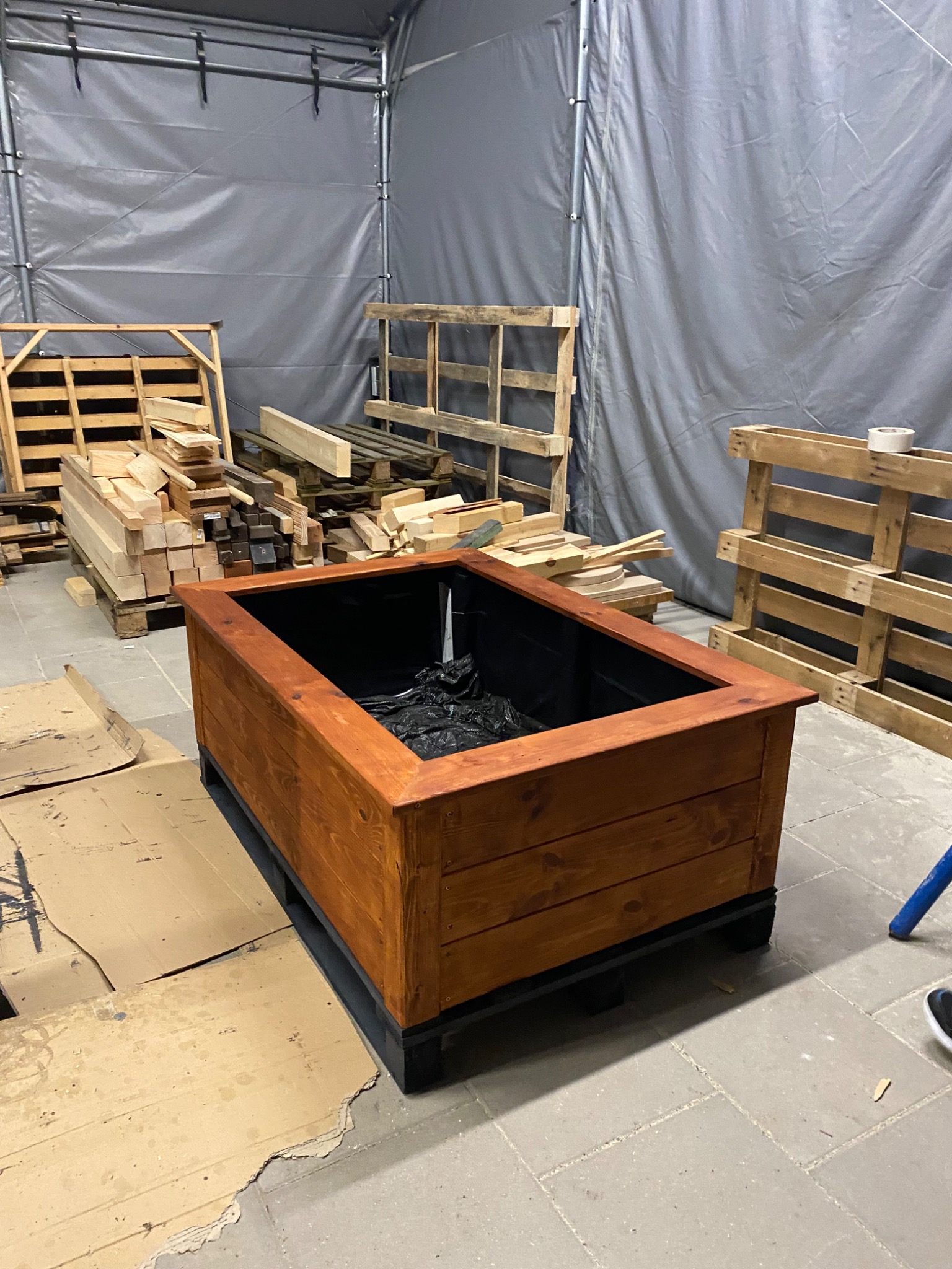 Drewniana skrzynia na rośliny, bejcowana na brązowo, z czarną wyściółką wewnątrz, stoi na palecie w warsztacie, w tle widoczne stosy drewna i palety.