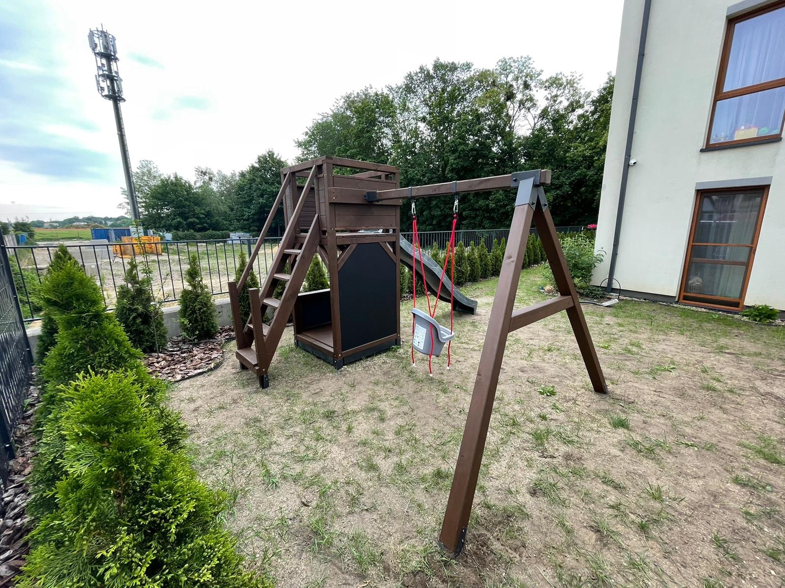 Drewniany plac zabaw z huśtawką kubełkową i zjeżdżalnią, umieszczony na trawiastym terenie w ogrodzie, z widoczną wieżą telekomunikacyjną w tle.