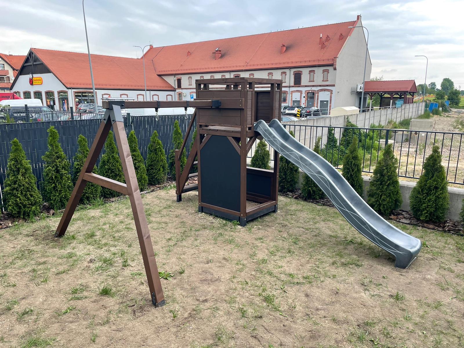 Drewniany plac zabaw z huśtawką i zjeżdżalnią na trawiastym podłożu, otoczony tujami i czarnym metalowym ogrodzeniem. W tle budynek z czerwoną dachówką.