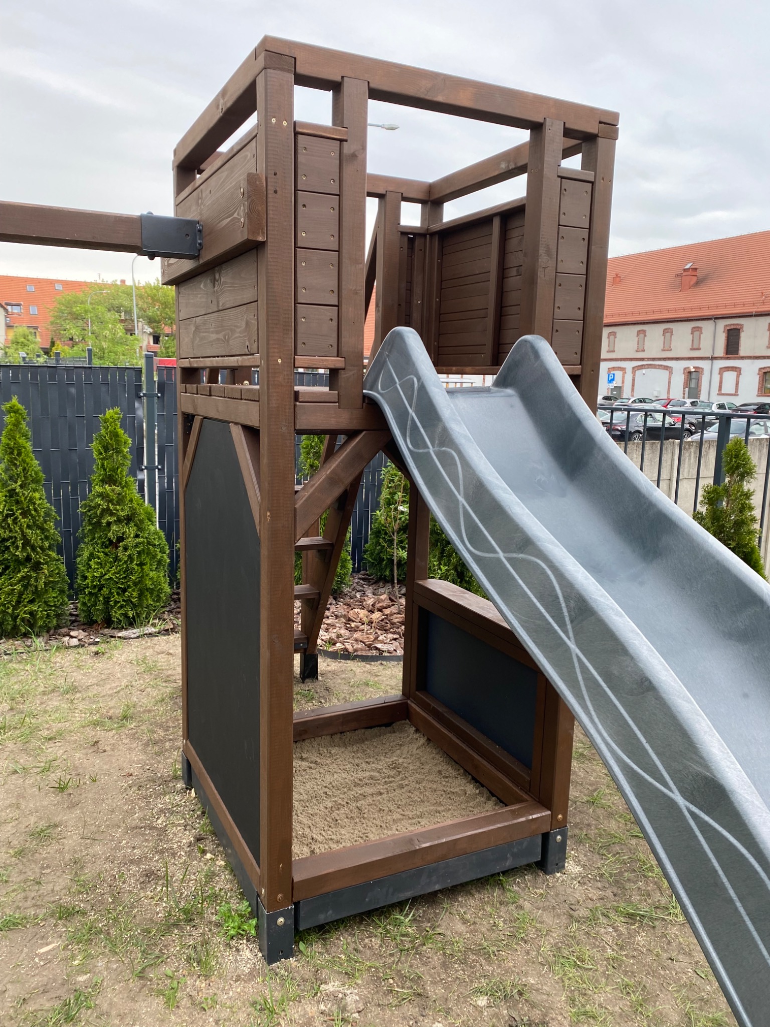 Drewniana konstrukcja placu zabaw z szarą zjeżdżalnią i tablicą, umieszczona na trawiastym terenie z widocznymi budynkami w tle.