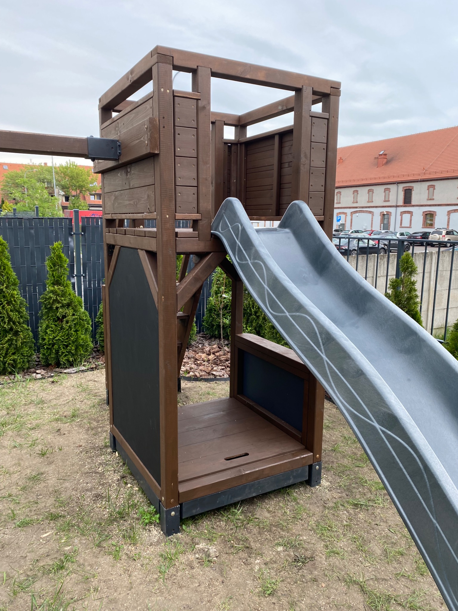 Drewniany plac zabaw z wieżą, zjeżdżalnią z dekoracyjnymi liniami i tablicą do pisania kredą, otoczony tujami i ogrodzeniem panelowym.